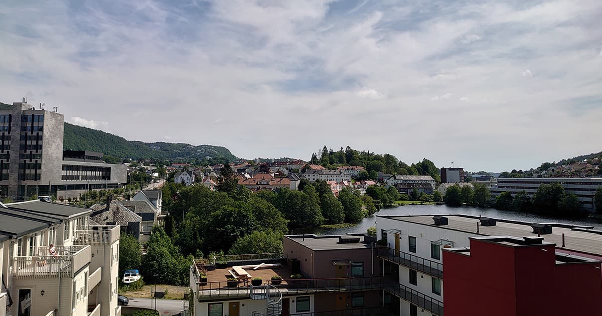 A view over Bergen, Norway, showing colourful buildings, lush green trees, and a calm river winding through the city with hills in the background under a partly cloudy sky.